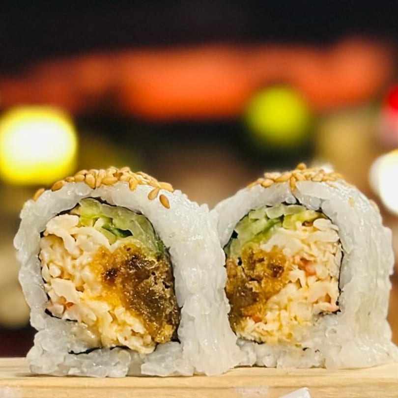 Two sushi rolls with rice, filling, and sesame seeds on a wooden surface with blurred lights.